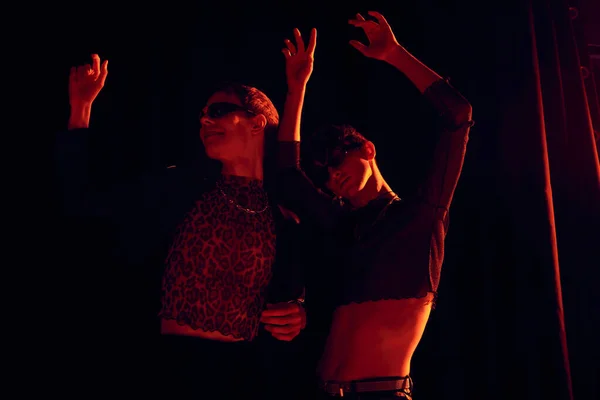 Amigos homossexuais na moda em óculos de sol e roupas de festa dançando enquanto celebra o mês de orgulho da comunidade lgbt no fundo preto com iluminação vermelha — Fotografia de Stock