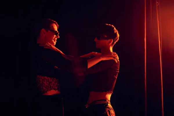Вид сбоку улыбающихся и трогательных молодых друзей-геев в солнцезащитных очках, обнимающих друг друга во время вечеринки и празднования месячника гордости на черном фоне с красной подсветкой — стоковое фото