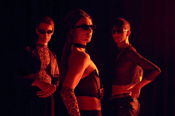 Grupo de la comunidad homosexual de moda en gafas de sol y trajes de fiesta posando juntos mientras se celebra lgbt mes aislado en negro con iluminación roja - foto de stock
