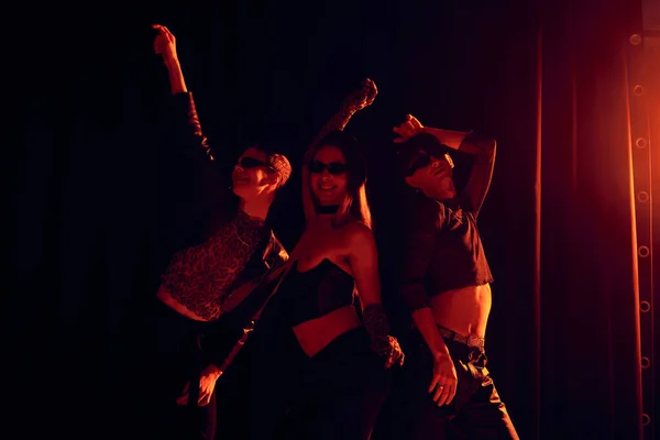 Groupe souriant d'amis homosexuels dans des vêtements élégants et des lunettes de soleil posant pendant la fête et la célébration du mois de la fierté lgbt sur fond noir avec éclairage rouge — Photo de stock