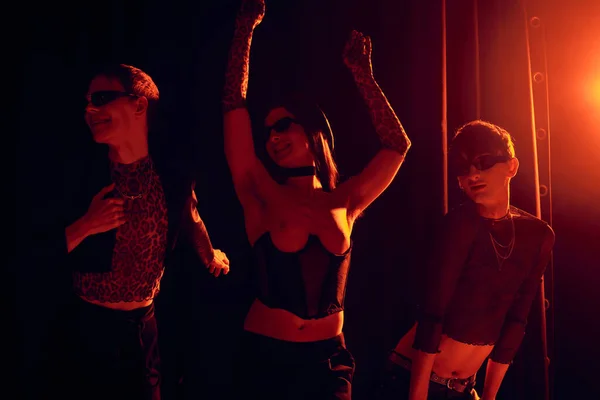 Aufgeregte Gruppe schwuler Freunde in Sonnenbrille und stilvoller Kleidung tanzen während der Party und lgbt Stolz Monatsfeier auf schwarzem Hintergrund mit rotem Licht — Stockfoto