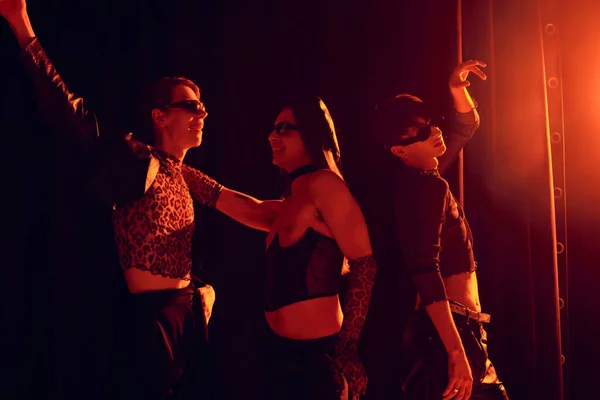 Sorrindo e pessoas homossexuais na moda em óculos de sol dançando durante a festa e lgbt orgulho mês celebração no fundo preto com iluminação vermelha — Fotografia de Stock