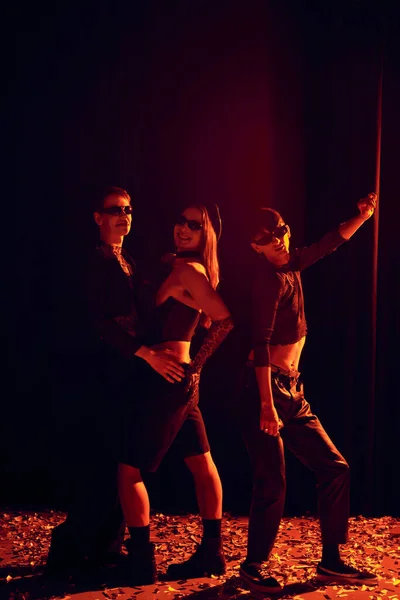 In voller Länge lächelnde und modische homosexuelle Menschen mit Sonnenbrille posieren auf Konfetti während der Party und lgbt Stolz Monatsfeier auf schwarzem Hintergrund mit rotem Licht — Stockfoto