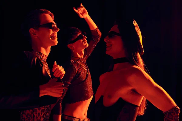 Вид сбоку улыбающихся и трогательных гомосексуалистов в солнцезащитных очках и праздничной одежде, танцующих во время празднования месячника гордости, изолированных черным с красным освещением — стоковое фото