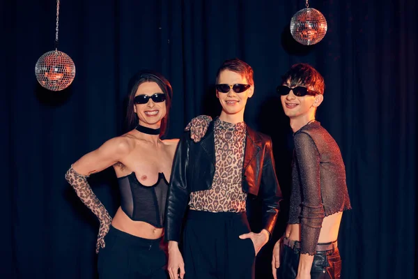 Cheerful and fashionable gay community in party outfits and sunglasses posing together near disco balls during pride month celebration on black background — Stock Photo