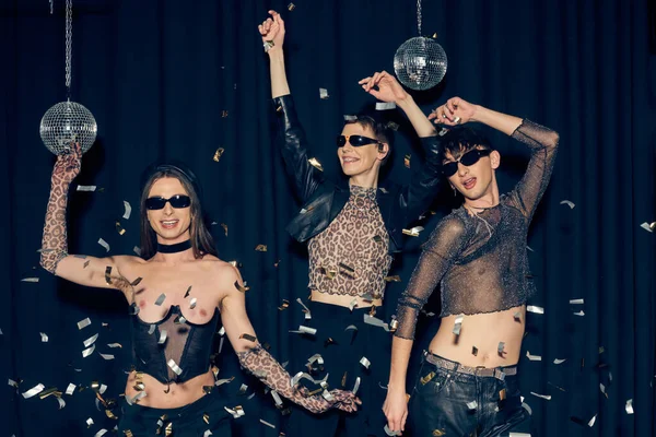 Fröhliche und modische schwule Freunde mit Sonnenbrille tanzen zusammen in der Nähe von Konfetti und Discokugeln während der Party und lgbt Monatsfeier auf schwarzem Hintergrund — Stockfoto