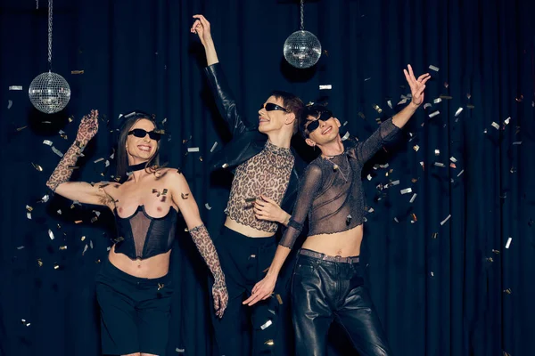 Smiling and stylish homosexual friends in sunglasses dancing during party near disco balls and confetti while celebrating lgbt pride month on black background — Stock Photo