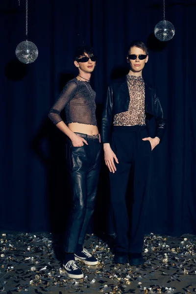 Fashionable queer friends in sunglasses posing while standing on confetti and posing with hands in pockets near disco balls during lgbt pride month party on black background — Stock Photo