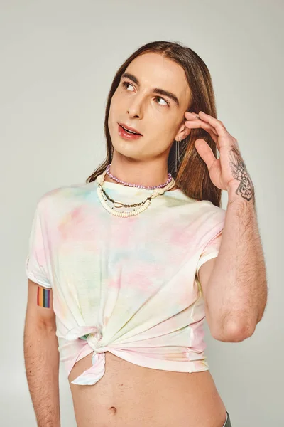 Мечтательный и длинноволосый гомосексуалист в футболке с галстуком, прикасающийся к волосам и отворачивающийся во время празднования месяца гордости, стоя изолированный на сером — стоковое фото