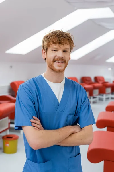 Trabalhador de saúde ruiva em uniforme azul de pé com braços dobrados e sorrindo para a câmera perto de cadeiras médicas confortáveis no centro de doação de sangue, fundo borrado — Fotografia de Stock