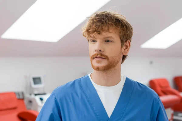 Молодой, рыжий и бородатый доктор в синей униформе стоит и смотрит в сторону размытых медицинских стульев в стерильной среде станции переливания крови — стоковое фото