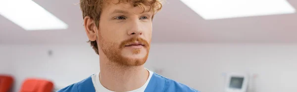 Young, redhead healthcare worker in blue uniform looking away in sterile environment of laboratory in blood donation center on blurred background, banner — Stock Photo