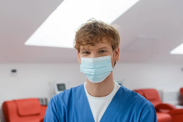 Redhead and young doctor in medical mask and blue uniform looking at camera in sterile environment of blood transfusion station near medical chairs on blurred background — Stock Photo
