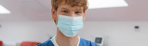 Young and redhead doctor with cheerful eyes expression, wearing medical mask and looking at camera in sterile environment of blood donation center on blurred background, banner — Stock Photo