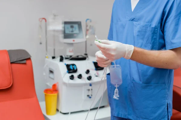 Vista recortada del médico en uniforme azul y guantes de látex estériles que sostienen el conjunto de transfusión de sangre cerca de equipo automatizado borroso y taza de plástico en el laboratorio - foto de stock