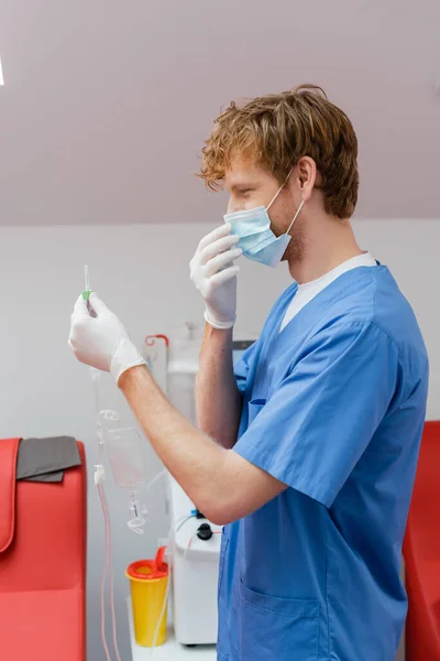 Вид сбоку молодой и рыжеволосый доктор в синей униформе, медицинской маске и латексных перчатках, держащих набор для переливания крови возле медицинских стульев в стерильной лаборатории — стоковое фото