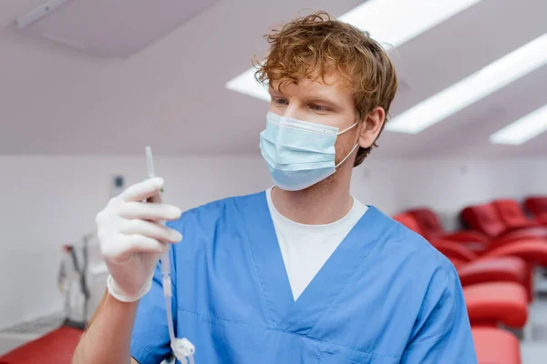 Young and redhead doctor in blue uniform, medical mask and latex glove holding blood transfusion set near ergonomic medical chairs on blurred background in hospital — Stock Photo