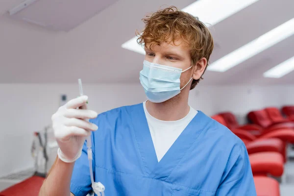 Médico joven y pelirrojo en uniforme azul, máscara médica y guante de látex que sostiene el conjunto de transfusión de sangre cerca de sillas médicas ergonómicas sobre fondo borroso en el hospital - foto de stock