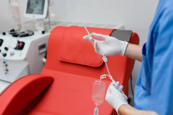 Частичный взгляд практикующего в стерильных латексных перчатках, держащих набор для переливания крови возле медицинского кресла с удобным эргономичным дизайном и современным оборудованием на размытом фоне в лаборатории — стоковое фото