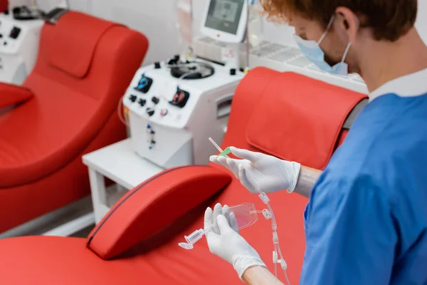 Jeune praticien en uniforme bleu et gants de latex stériles tenant ensemble de transfusion près de l'équipement automatisé et des chaises médicales dans le centre de don de sang, premier plan flou — Photo de stock