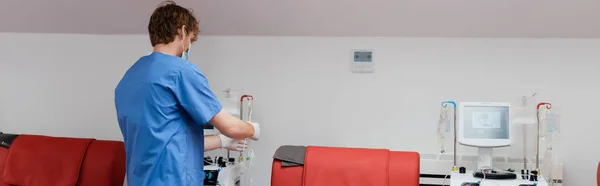 Вид ззаду на рудого лікаря в блакитній формі, медична маска і латексні рукавички, що працюють з обладнанням для переливання крові біля червоних медичних стільців і крапельних стендів в медичній лабораторії, банер — стокове фото