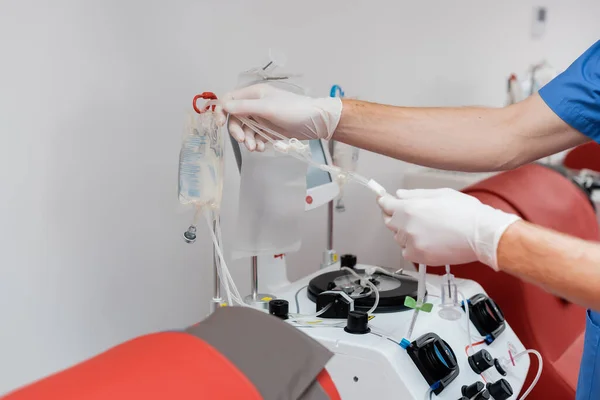 Partial view of doctor in sterile latex gloves assembling blood transfusion set on drip stand with infusion bags near automated equipment and red medical chairs in medical laboratory — Stock Photo