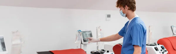 Вид збоку лікаря в медичній масці та синій формі, що стоїть біля сенсорного екрану та операційної машини переливання крові в стерильній медичній лабораторії, банер — стокове фото