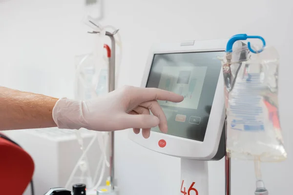 Vista parcial del trabajador sanitario en guante de látex operando una moderna máquina de transfusión automatizada con pantalla táctil cerca de soportes de goteo y bolsas de infusión en laboratorio médico - foto de stock
