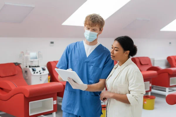 Young redhead doctor in blue uniform, medical mask and latex gloves showing digital tablet to multiracial woman near blurred medical chairs in blood donation center — Stock Photo