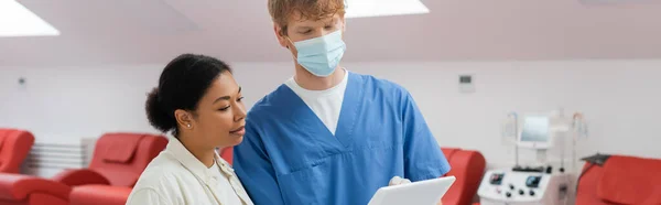 Multirassische Frau und rothaarige Ärztin in blauer Uniform und medizinischer Maske mit Blick auf digitales Tablet in der Nähe von Medizinstühlen und Bluttransfusionsgerät in der Klinik, Banner — Stockfoto