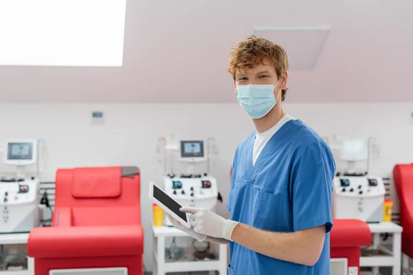 Trabalhador de saúde ruiva em uniforme azul, máscara médica e luvas de látex olhando para a câmera perto de cadeiras médicas e máquinas de transfusão em fundo turvo no centro de doação de sangue — Fotografia de Stock