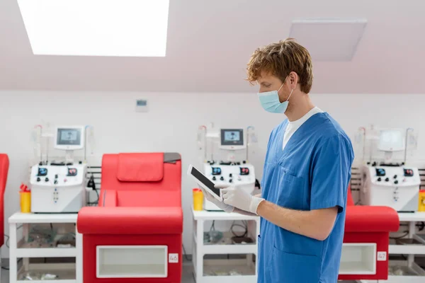 Médico pelirrojo en máscara médica, uniforme azul y guantes de látex usando tableta digital con pantalla en blanco cerca de sillas médicas borrosas y máquinas automatizadas de transfusión en el centro de donación de sangre - foto de stock