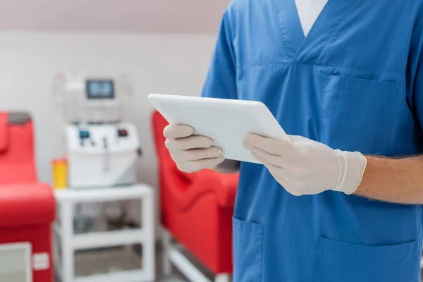 Visão recortada do médico de uniforme azul e luvas de látex segurando tablet digital perto de cadeiras médicas borradas e máquina de transfusão automatizada no centro de doação de sangue — Fotografia de Stock