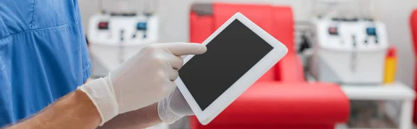Partial view of doctor in blue uniform and latex gloves pointing at digital tablet with blank screen near blurred medical chair and transfusion machines in laboratory, banner — Stock Photo