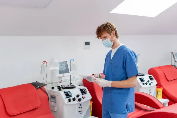 Рыжеволосый врач в синей форме, медицинской маске и латексных перчатках смотрит на цифровой планшет рядом с медицинскими стульями и автоматизированными машинами для переливания крови в центре донорства — стоковое фото