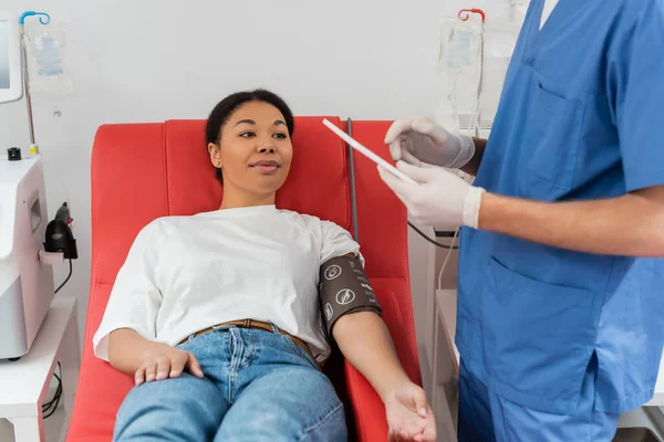 Vielrassige Frau in Blutdruckmanschette sitzt auf dem Behandlungsstuhl in der Nähe automatisierter Transfusionsmaschinen und schaut den Arzt in blauer Uniform und Latexhandschuhen mit digitalem Tablet in der Klinik an — Stockfoto