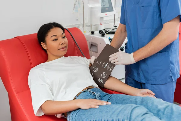 Gesundheitshelfer in blauer Uniform und Latexhandschuhen legt Blutdruckmanschette auf den Arm einer multiethnischen Frau, die auf einem bequemen medizinischen Stuhl in der Nähe eines automatischen Transfusionsautomaten im Krankenhaus sitzt — Stockfoto