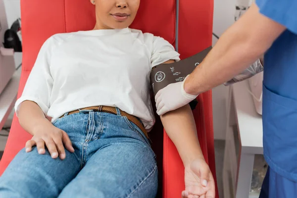 Частковий вигляд лікаря в латексних рукавичках, що регулює тиск манжети на багаторасову жінку, що сидить на зручному медичному кріслі в станції переливання крові, догляд за пацієнтами — стокове фото