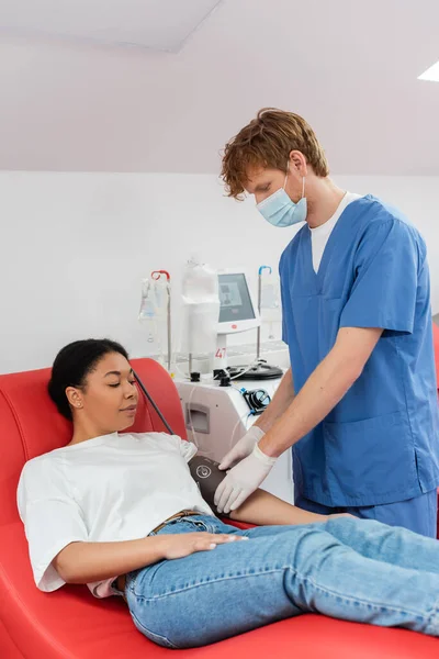 Redhead healthcare worker in blue uniform, medical mask and latex gloves adjusting blood pressure cuff on arm of multiracial woman sitting on medical chair near transfusion machine, patient care — Stock Photo