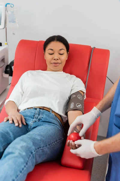 Gesundheitshelfer in Latex-Handschuhen gibt Gummiball an multirassische Frau in Blutdruckmanschette, die auf bequemen medizinischen Stuhl in zeitgenössischer Klinik sitzt, Patientenversorgung — Stockfoto