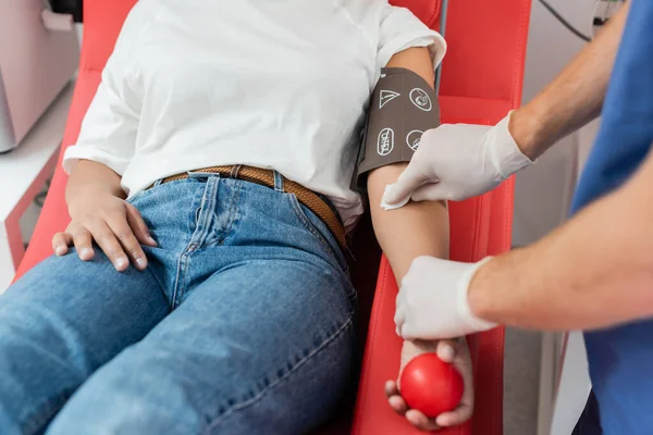 Teilansicht des Arztes in Latex-Handschuhen, der den Arm einer multirassischen Frau in Blutdruckmanschette abwischt, die auf dem Behandlungsstuhl sitzt und Gummiball im Labor hält, Patientenversorgung — Stockfoto