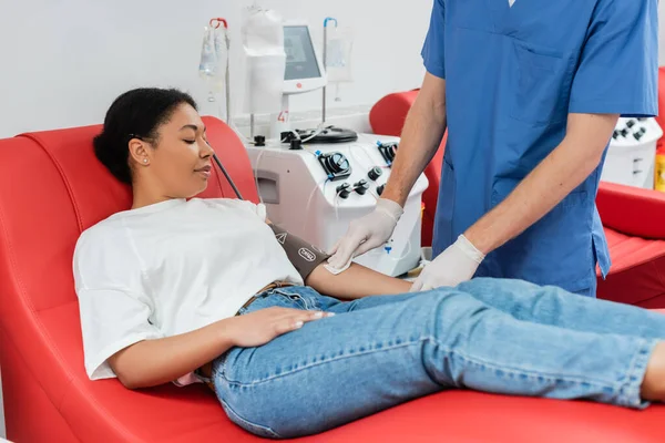 Healthcare worker in blue uniform and latex gloves holding alcohol pad and wiping arm of multiracial woman in blood pressure cuff sitting on medical chair near transfusion machine — Stock Photo