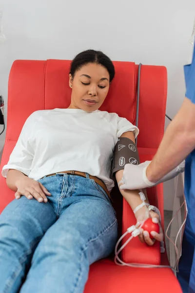 Trabajador de la salud que conecta el equipo de transfusión de sangre a la mujer multirracial sentada en una silla médica cómoda en el manguito de presión arterial y apretando la bola de goma - foto de stock