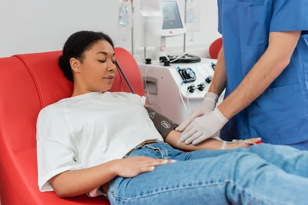 Врач в латексных перчатках приклеивает пластырь к руке многорасовой женщины, сидящей на медицинском стуле и сдающей кровь возле автоматизированной машины для переливания — стоковое фото