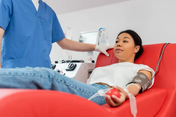 Medico in uniforme blu in piedi vicino alla donna multirazziale seduto su una comoda sedia medica vicino alla macchina trasfusione mentre dona sangue in laboratorio medico — Foto stock