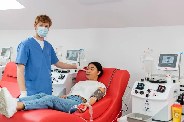 Médecin en masque médical, uniforme bleu et gants en latex regardant la caméra près des machines à transfusion et femme multiraciale assise sur une chaise médicale tout en faisant un don de sang à la clinique — Photo de stock
