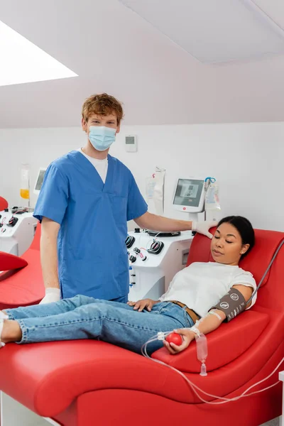 Médecin rousse en masque médical et uniforme bleu regardant la caméra près des machines de transfusion automatisées et multiracial assis sur une chaise médicale et faisant don de sang en laboratoire — Photo de stock