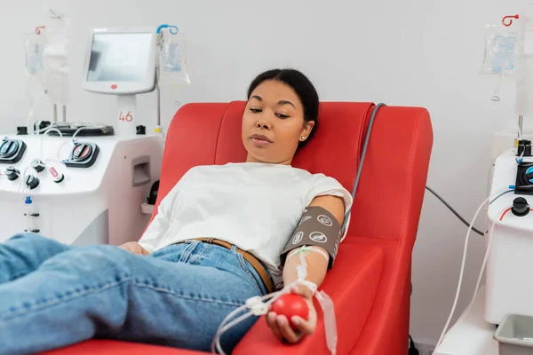 Mulher multirracial com conjunto de transfusão e bola de borracha sentada em cadeira médica ergonômica perto de máquinas de transfusão e doando sangue no hospital — Fotografia de Stock