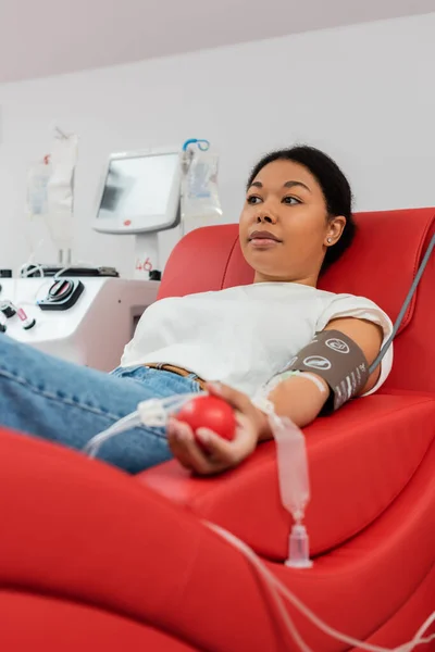 Mulher multirracial com conjunto de transfusão e bola de borracha médica sentado em cadeira médica confortável perto de equipamentos automatizados e doando sangue em laboratório — Fotografia de Stock
