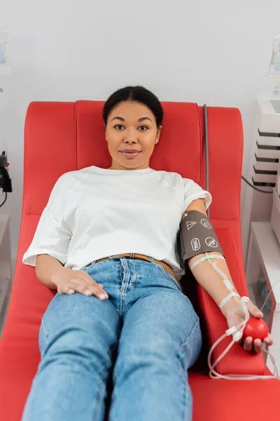 Положительная многорасовая женщина с наручниками давления, набор для переливания крови и медицинский резиновый мяч сидя на медицинском стуле и глядя на камеру во время сдачи крови в клинике — стоковое фото